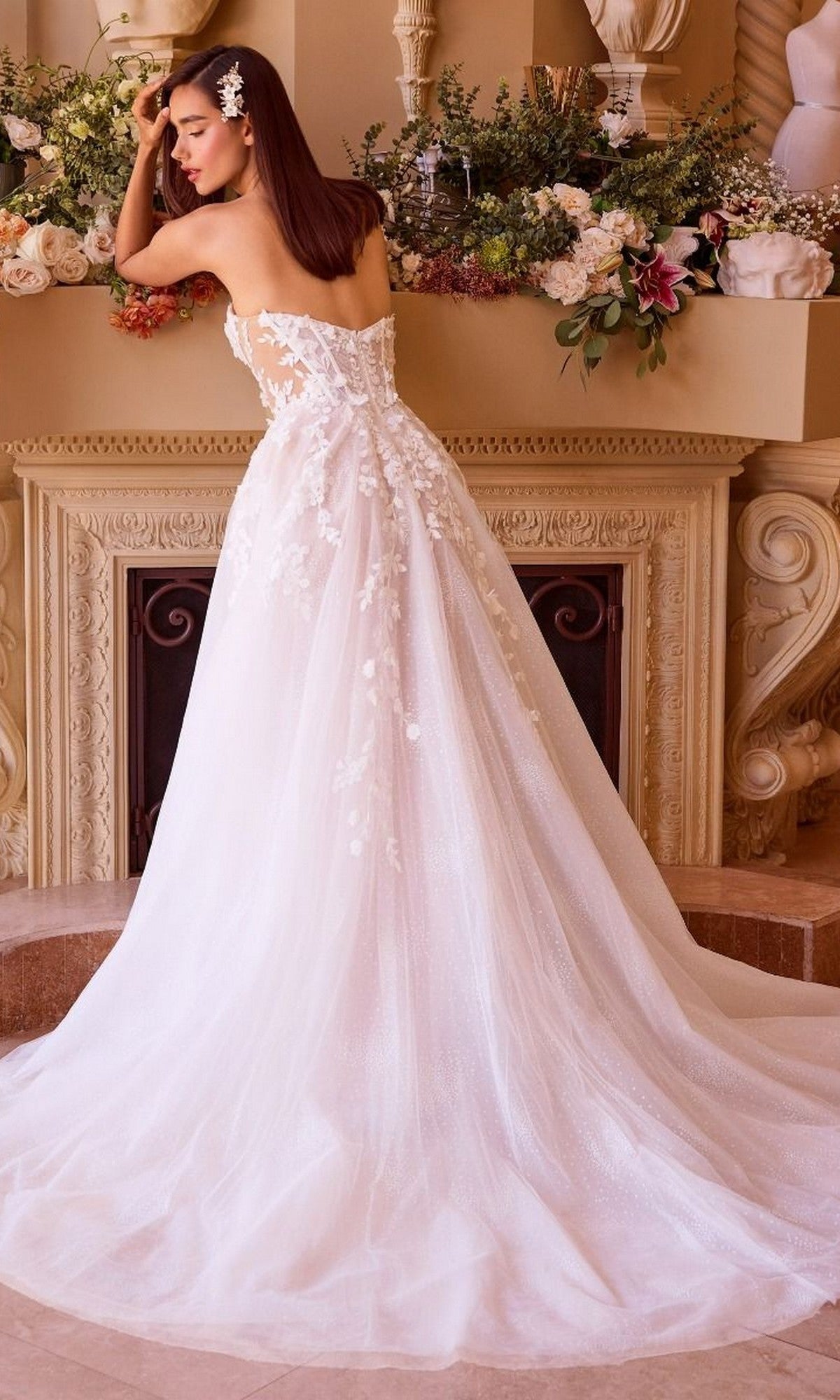 A woman in the Andrea and Leo WL032 Long Wedding Dress, featuring a strapless white lace and tulle A-line skirt, stands with her back to the camera, leaning on a flower-adorned mantel for a romantic touch.